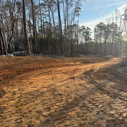 Cleared dirt road curving through a pine forest, with a white truck parked on the left.