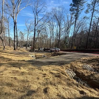 Leafless wooded clearing with a dirt path, parked vehicles, and a creek in the foreground under a blue sky