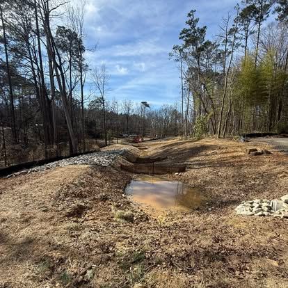 Rural dirt road with muddy puddle, leafless trees, and piles of earth under a partly cloudy sky