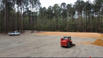 Construction site with gravel lot, orange skid steer, white pickup, and pine trees in the background