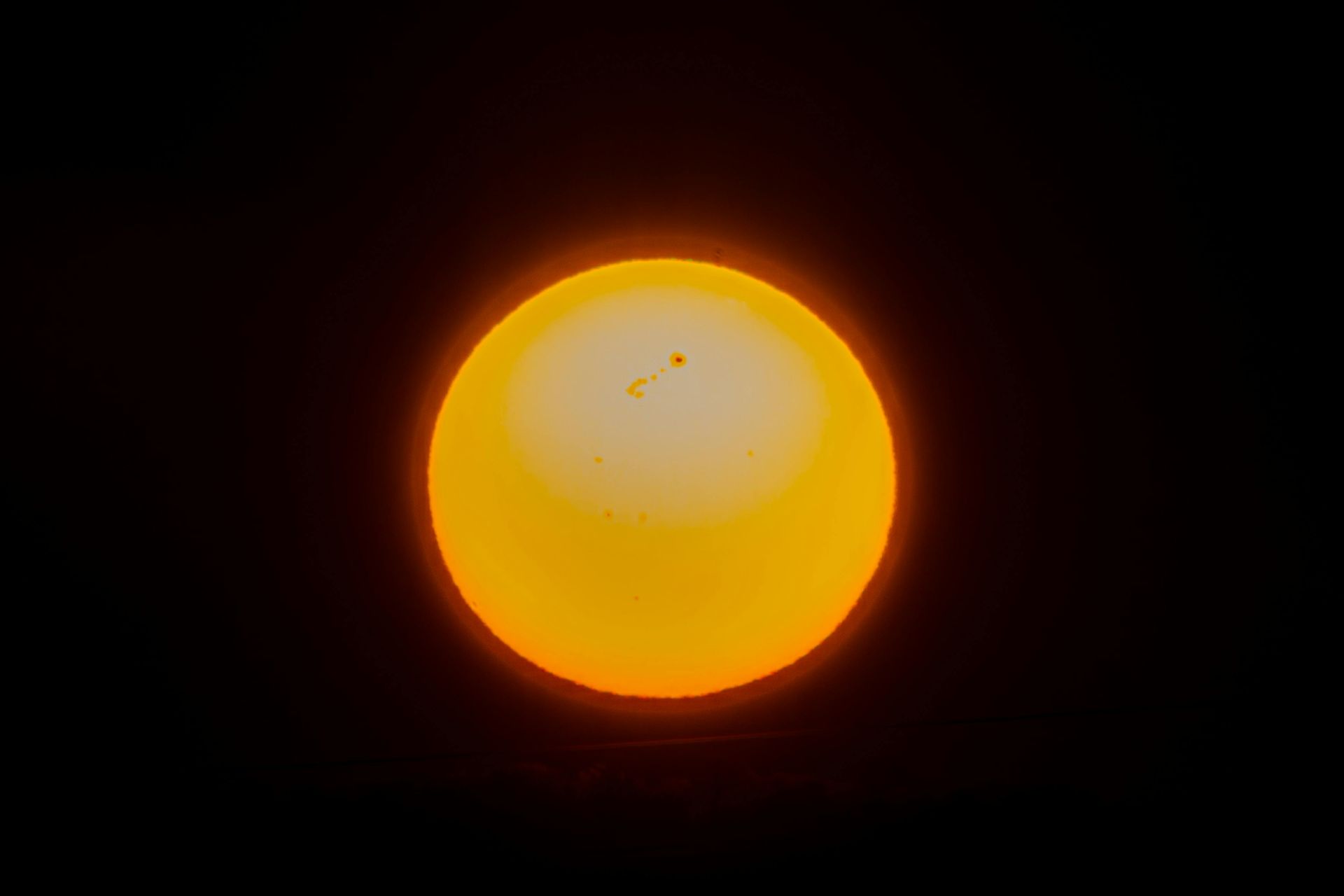 A bright, glowing yellow sun set against a solid black background, featuring small dark sunspots near its center.