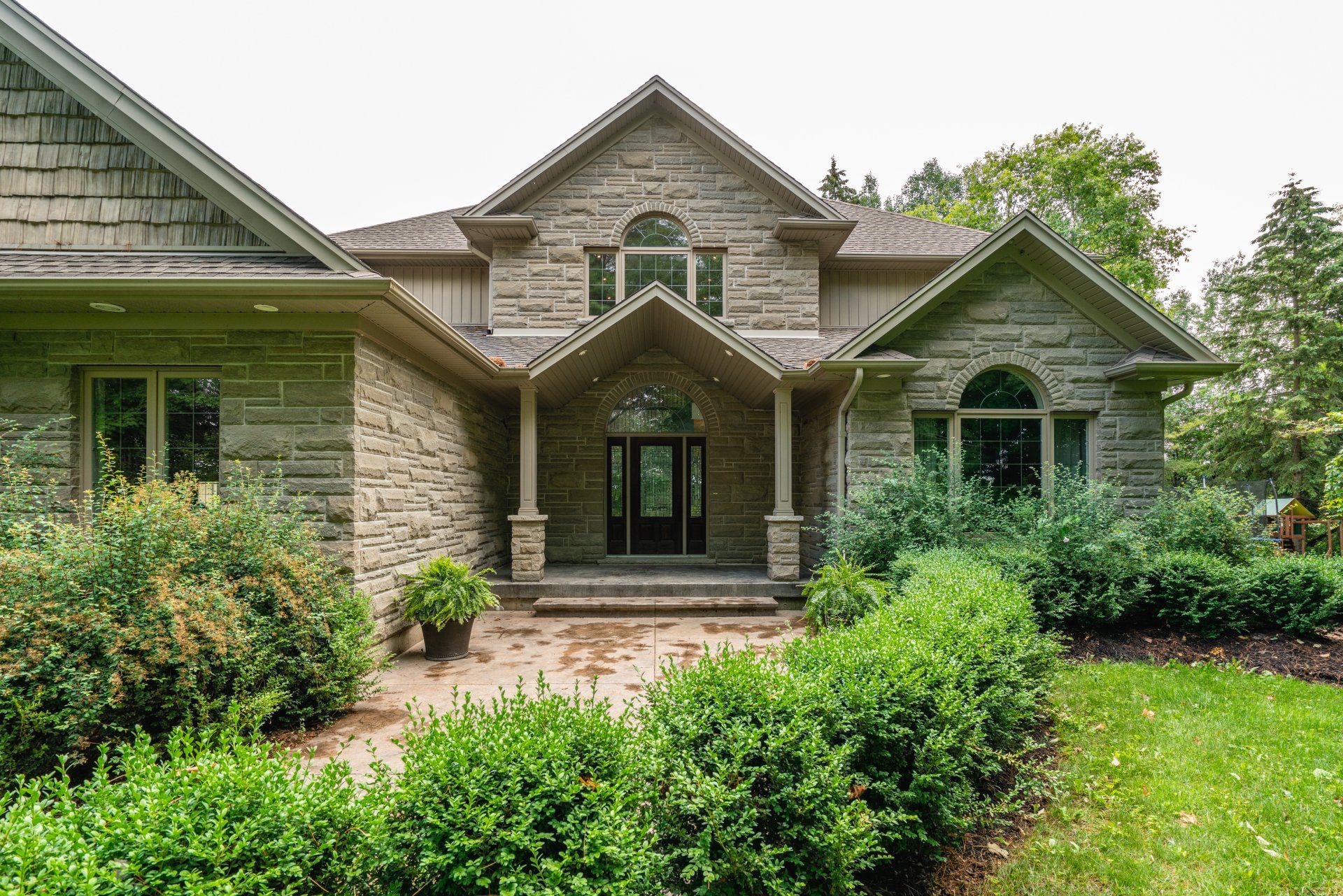 The front of a large stone house with a porch and bushes in front of it.