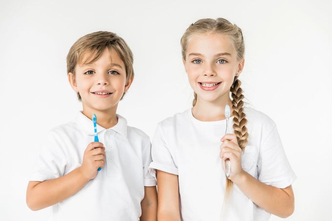 Due bambini con indosso camicie bianche, che tengono in mano degli spazzolini da denti e sorridono su uno sfondo bianco.