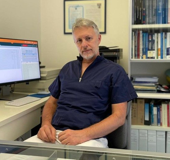 Uomo seduto a una scrivania in ufficio, con indosso una divisa blu scuro, con scaffali e un computer nelle vicinanze.