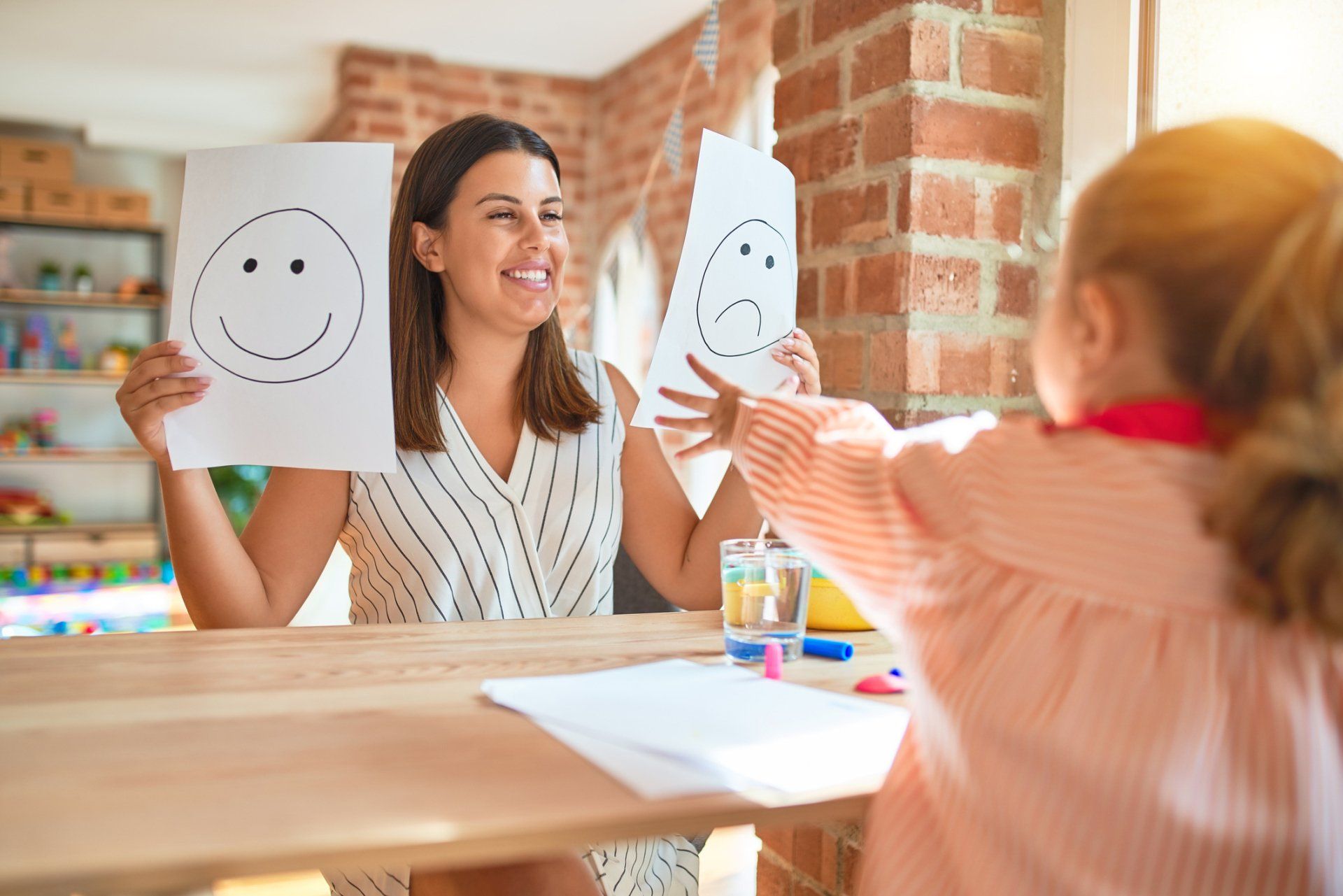 Teacher and child holding smiley and sad face cards across a table in a bright classroom