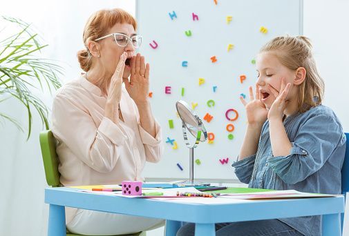 Teacher and child doing a face exercise at a table with a mirror and colorful learning materials