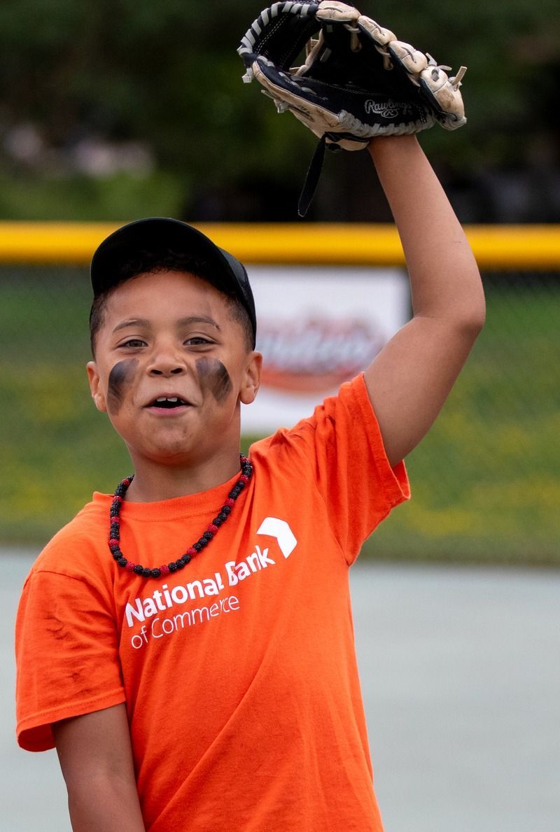 Boy in orange shirt with baseball glove raised, black eye paint, outdoors.