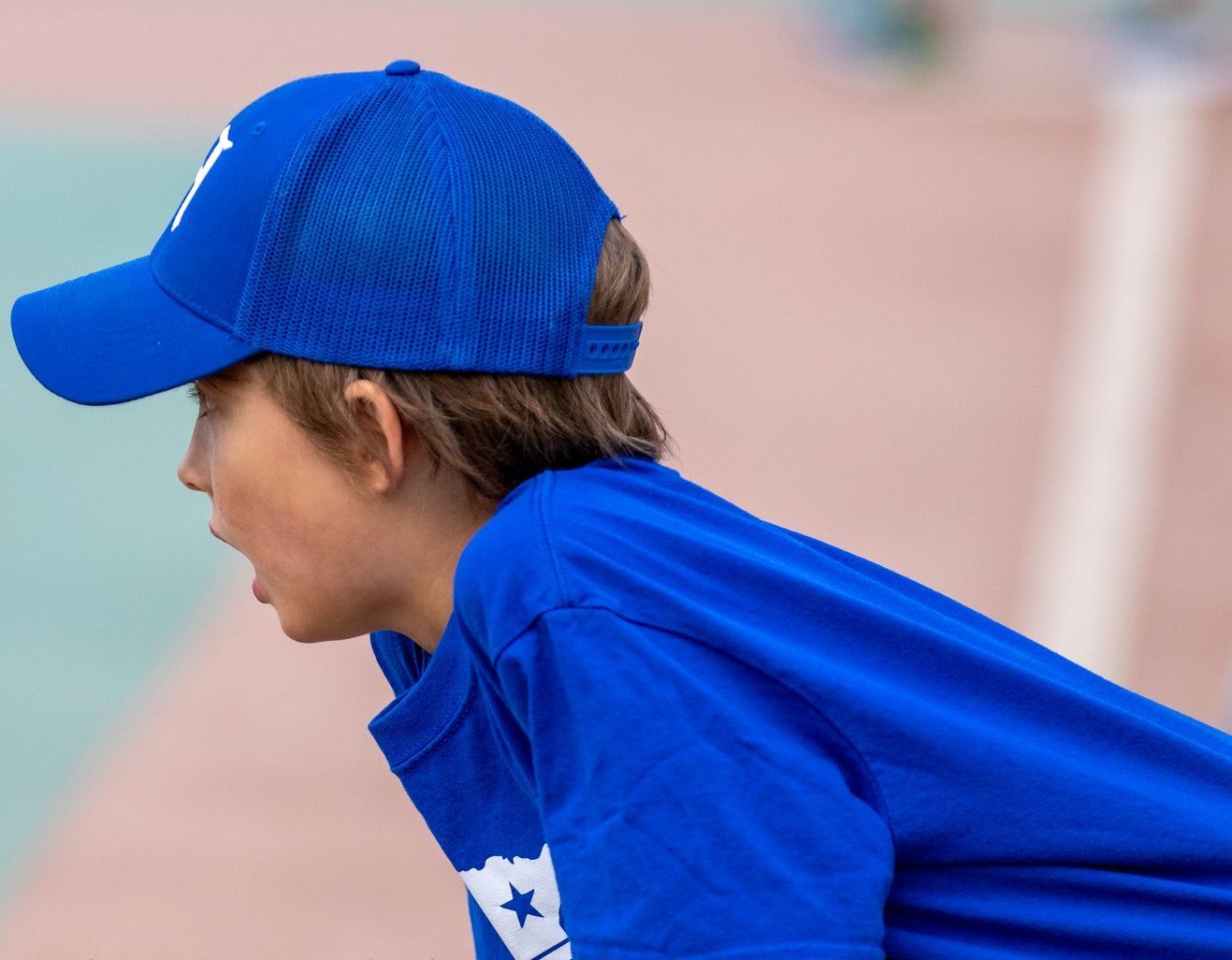 Boy in blue cap and shirt, mouth open, looking left, on a court or field, presumably cheering.