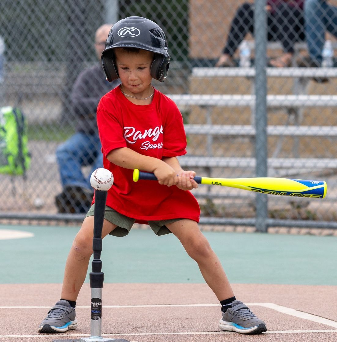 Young batter in red shirt and helmet swinging at a ball on a tee at a baseball field.