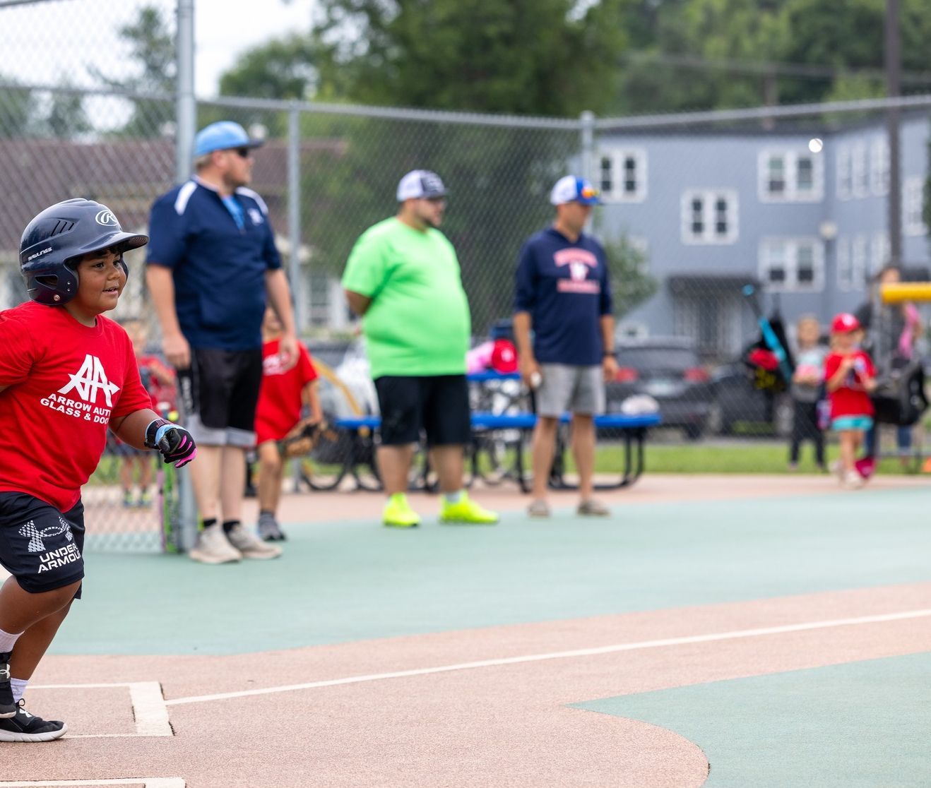 Child running toward base in a baseball game. Adults watch in the background near a fence.