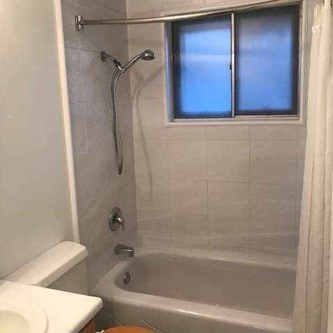 Small bathroom with bathtub, showerhead, toilet, and frosted window with blue light outside