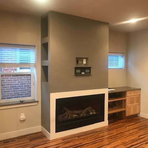 Fireplace with built-in shelves, gray wall, white trim, wooden floor, windows, and a small wooden cabinet.
