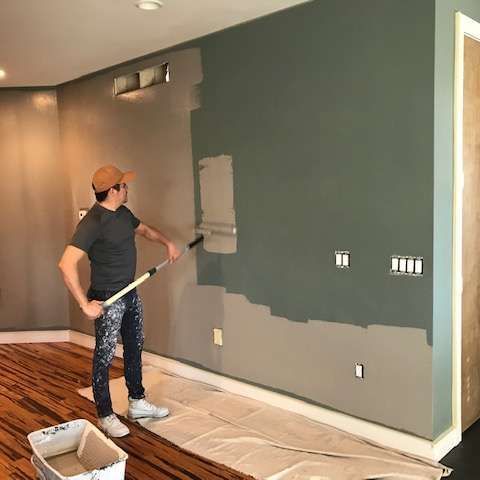 Person painting a wall gray in a room with hardwood floors and drop cloths
