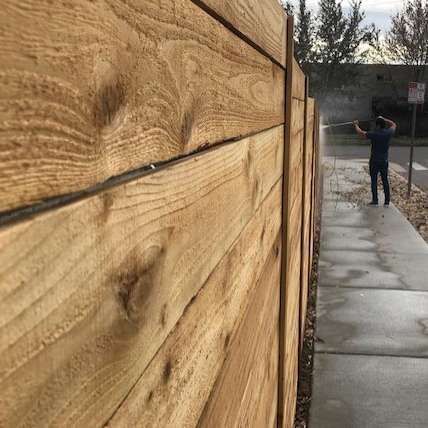 Close-up of a wooden fence along a wet concrete path, with a person spraying water in the distance.