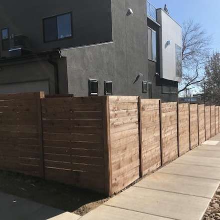Dark house beside a sidewalk, with a tall wooden fence along the street under a clear blue sky
