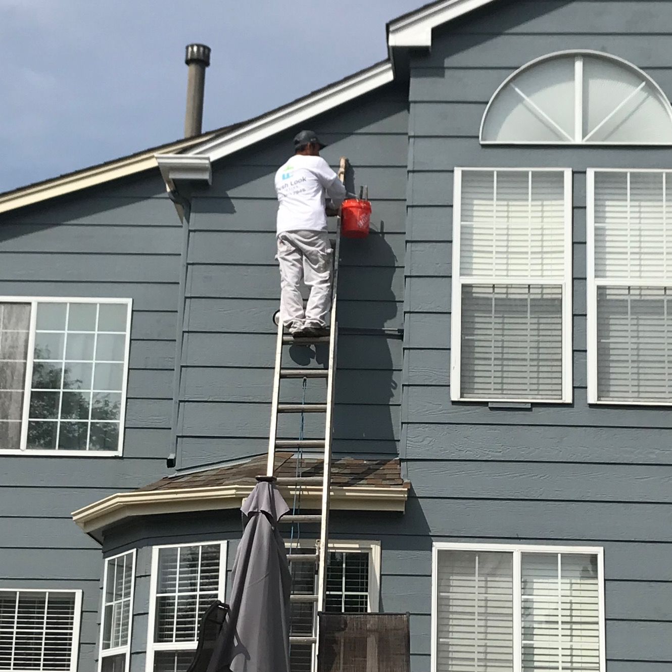 Person on a ladder painting a blue house, holding a red bucket, against a cloudy sky.