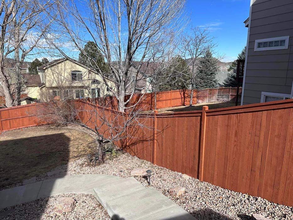 Red stained wooden fence surrounds a backyard with concrete path and gravel.