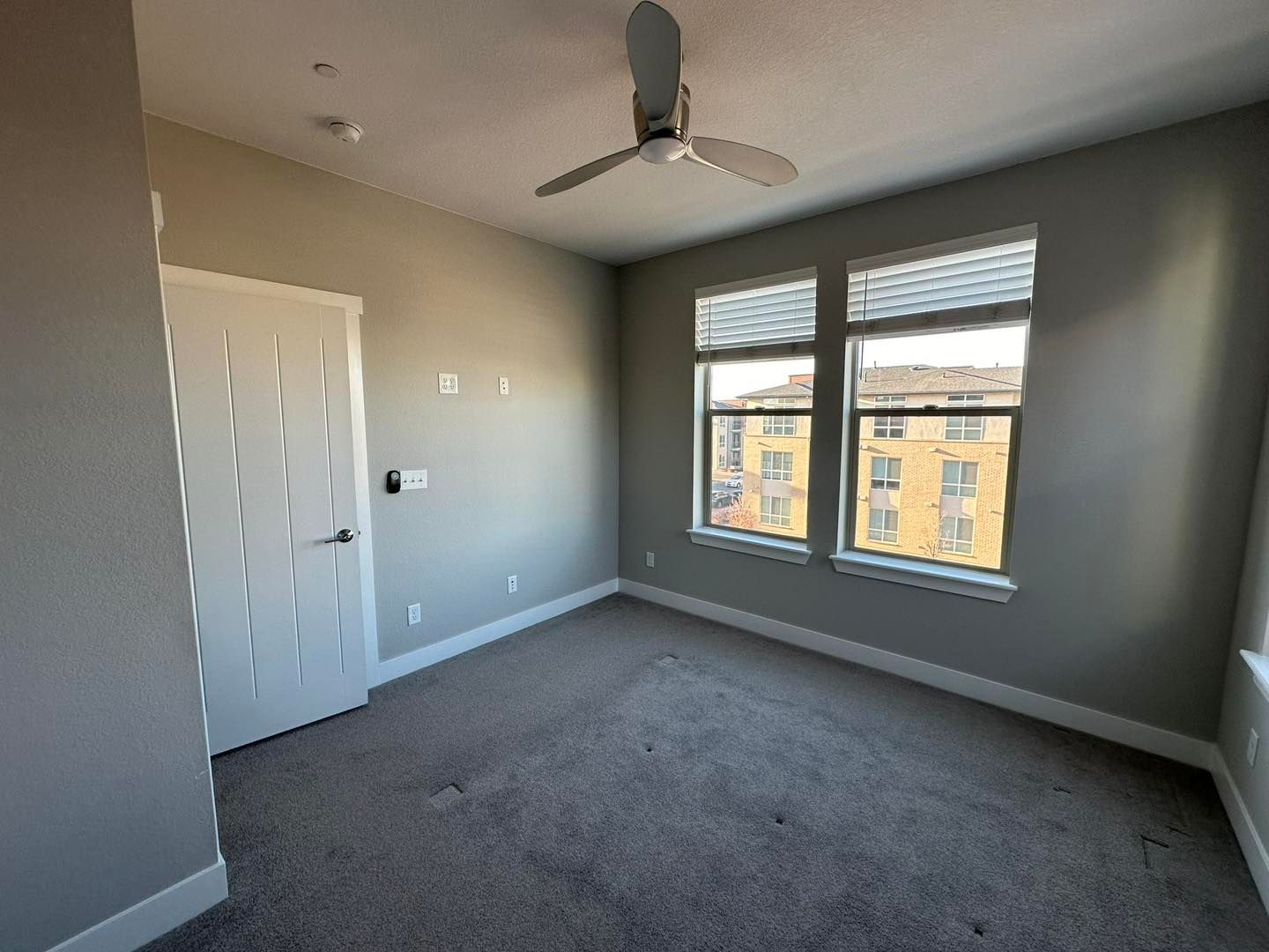 Empty bedroom with gray carpet, light walls, ceiling fan, and two windows above a radiator.
