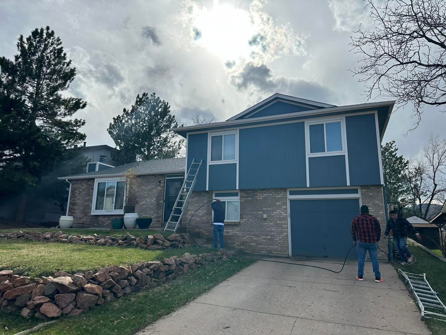Two-story blue house with stone exterior and driveway; two people near garage ladder and ladder on ground