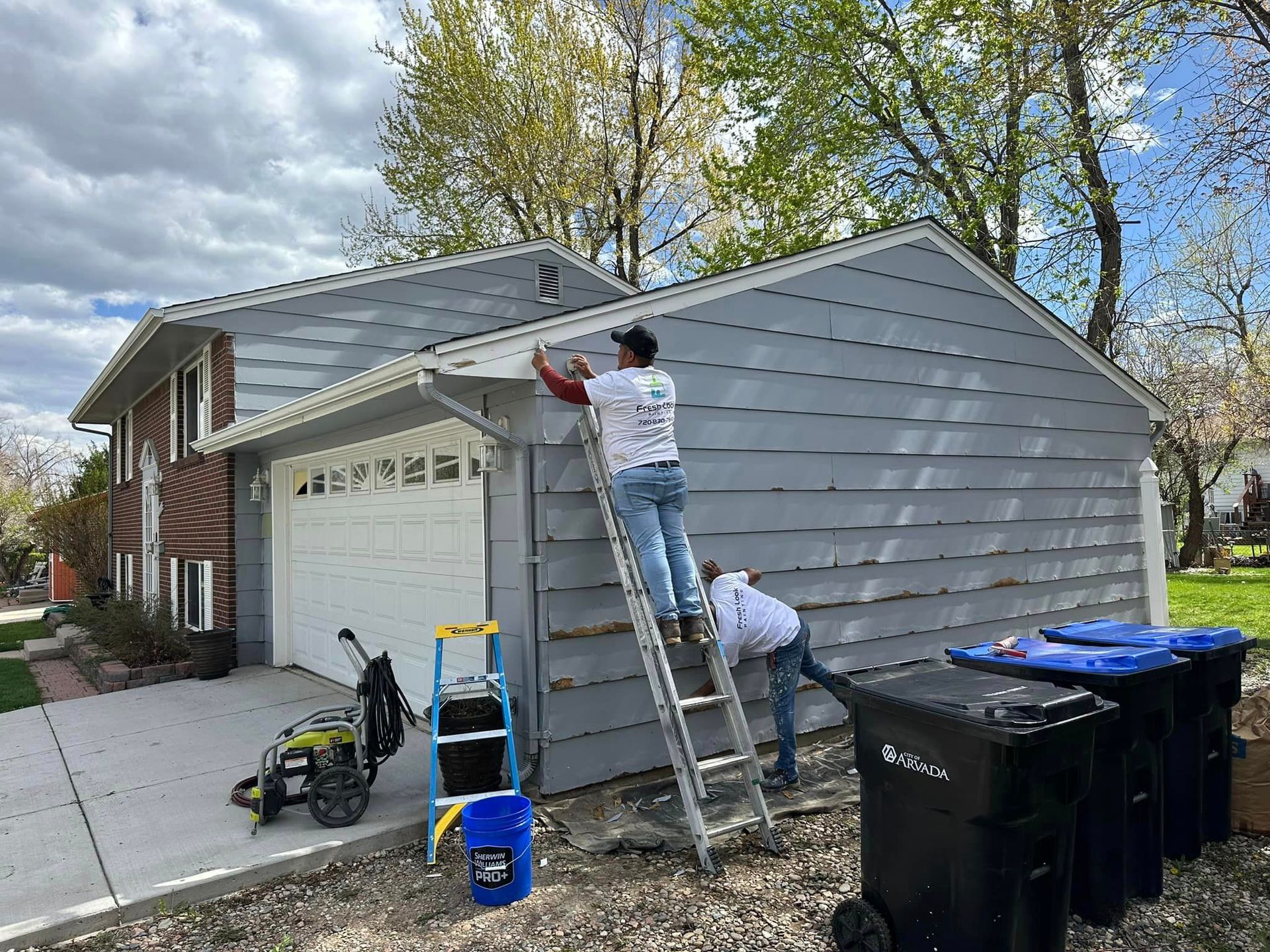 Two people painting a gray house. One on a ladder, the other below. Trash bins and a power washer are present.