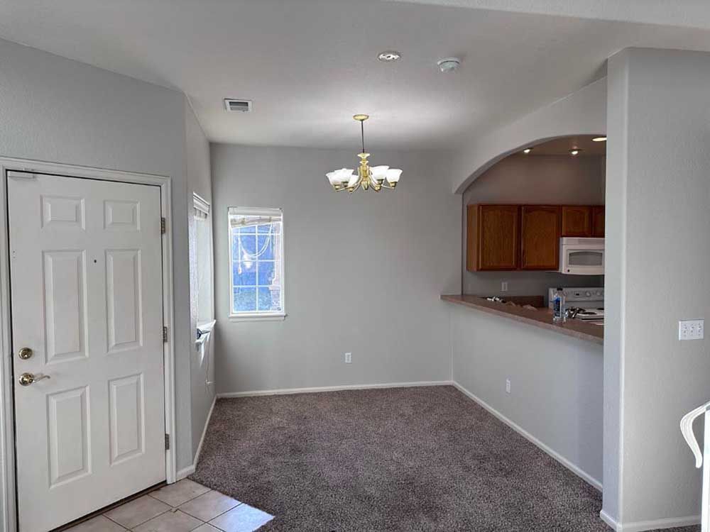Empty living room with gray carpet, white walls, front door, chandelier, and open kitchen pass-through