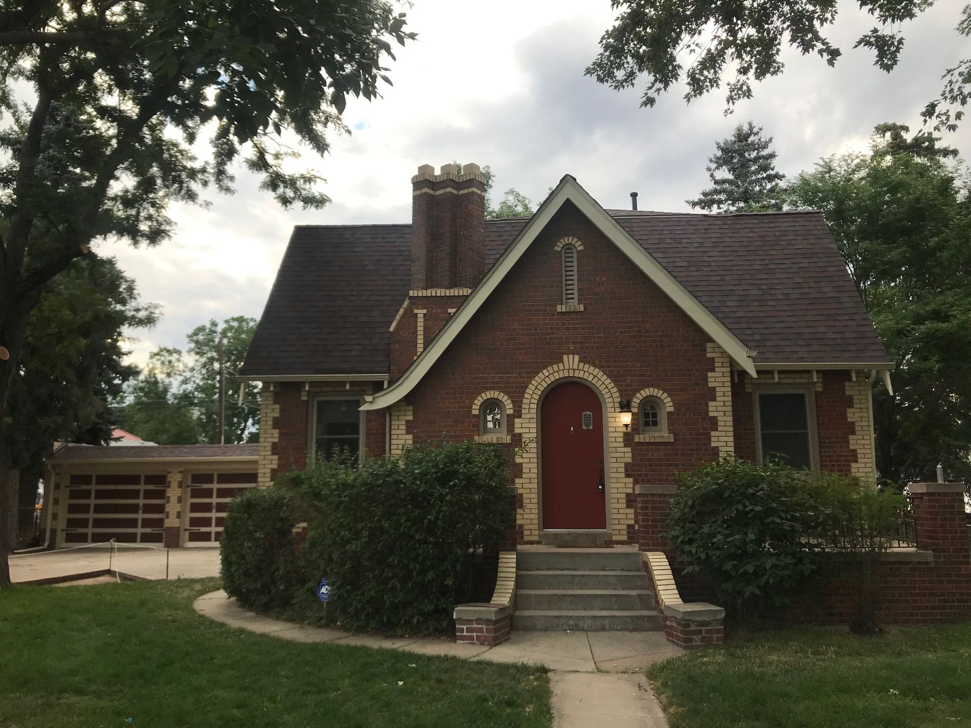 Red brick house with red door, small windows, and dark roof. Green bushes and a garage are visible.