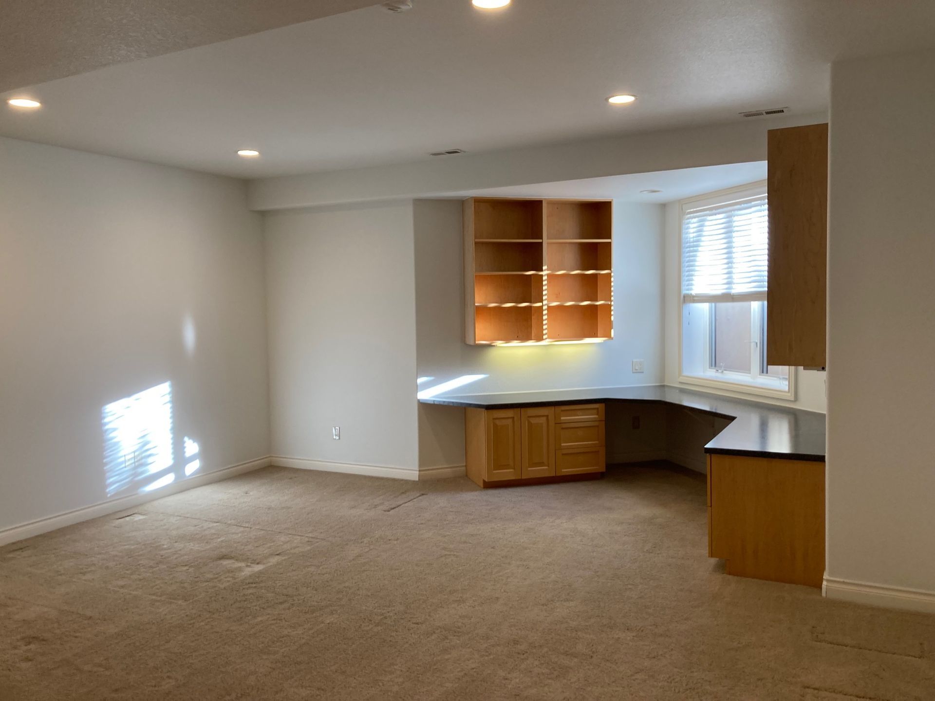 Empty room with built-in desk, cabinets, and a window. Beige carpet, white walls.