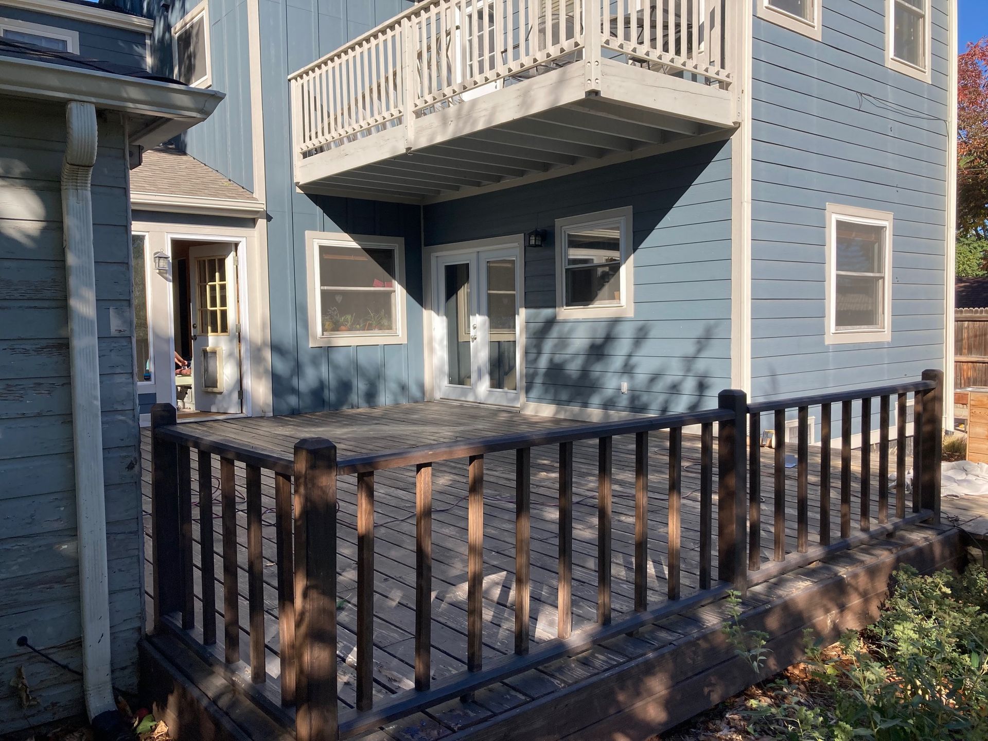 Blue house with wooden deck, balcony. Doors, windows visible. Deck has dark railing.