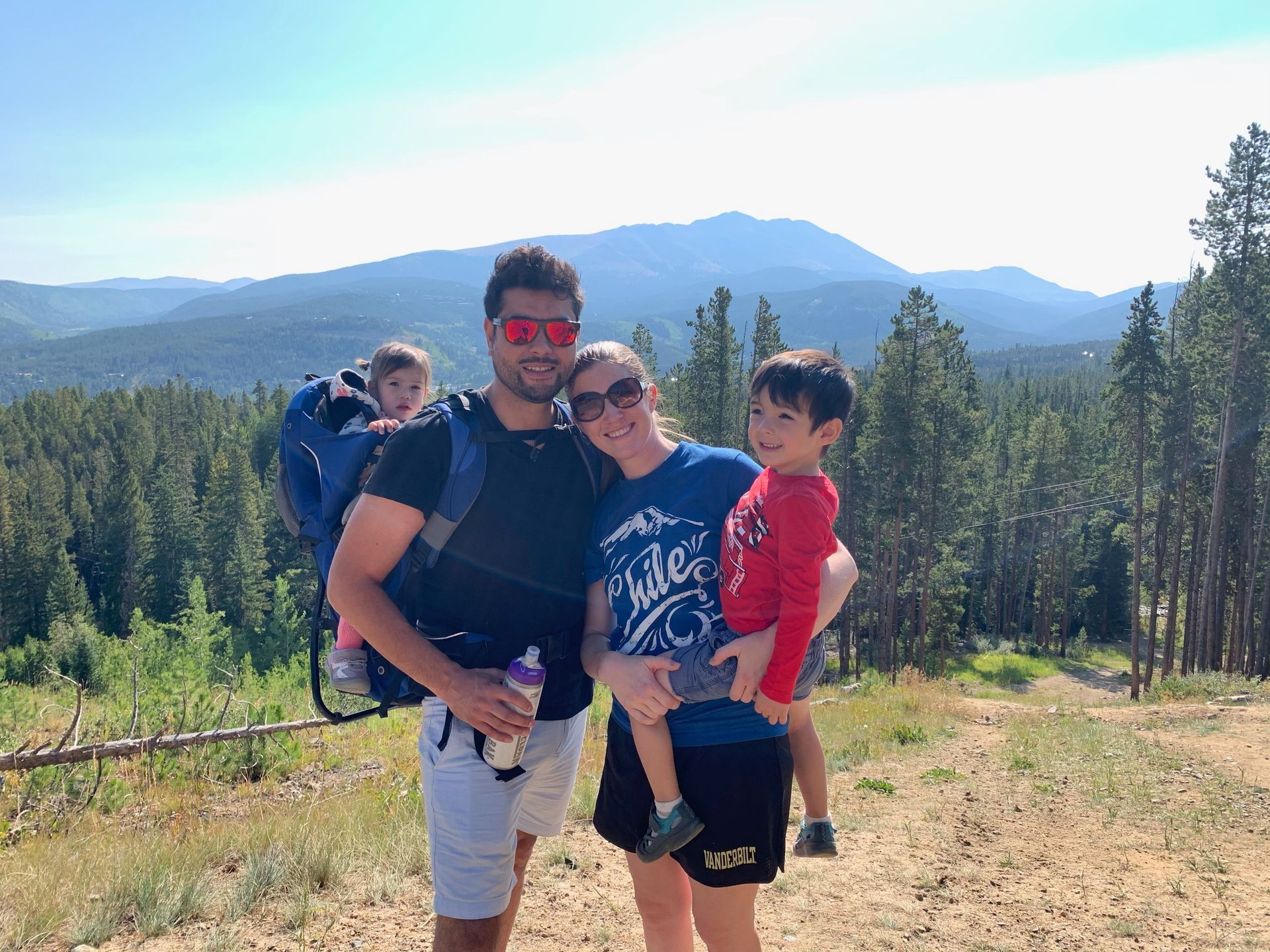 Family of four smiling on a hillside overlooking mountains; man carries child, woman holds another child.