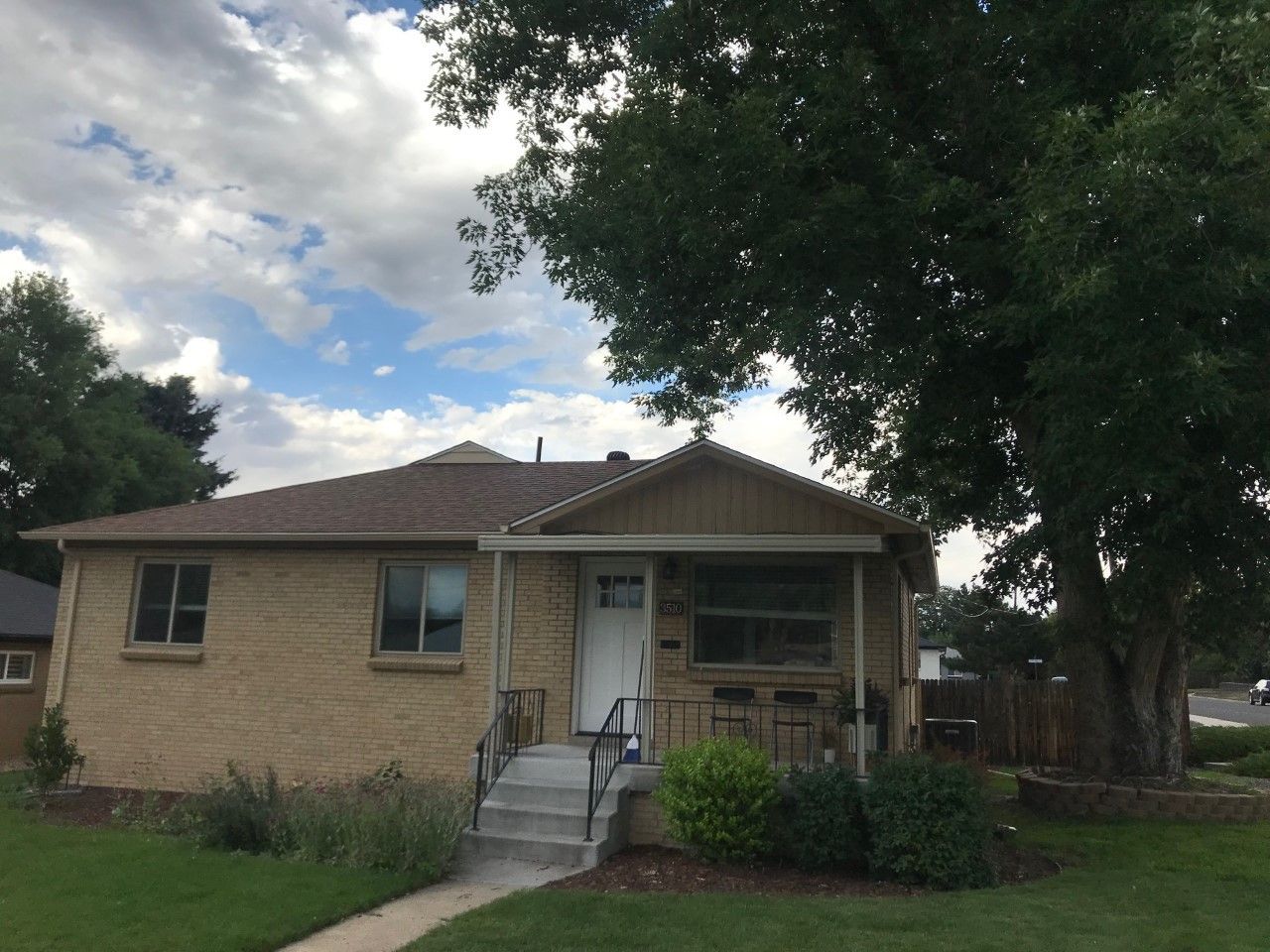 Beige brick house with small porch, cloudy sky, and large tree.