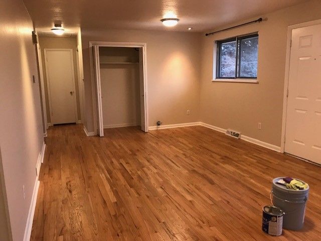Empty beige room with hardwood floors, ceiling lights, a window, and paint supplies on the floor