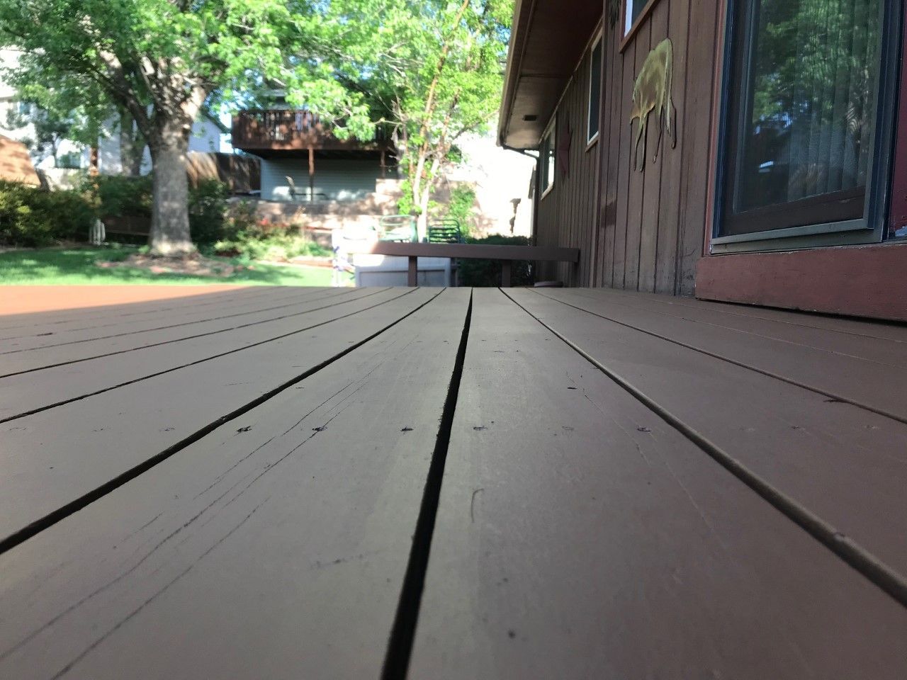 Low-angle view of a brown wooden deck with cracks, next to a brown house; trees and another structure are in the background.