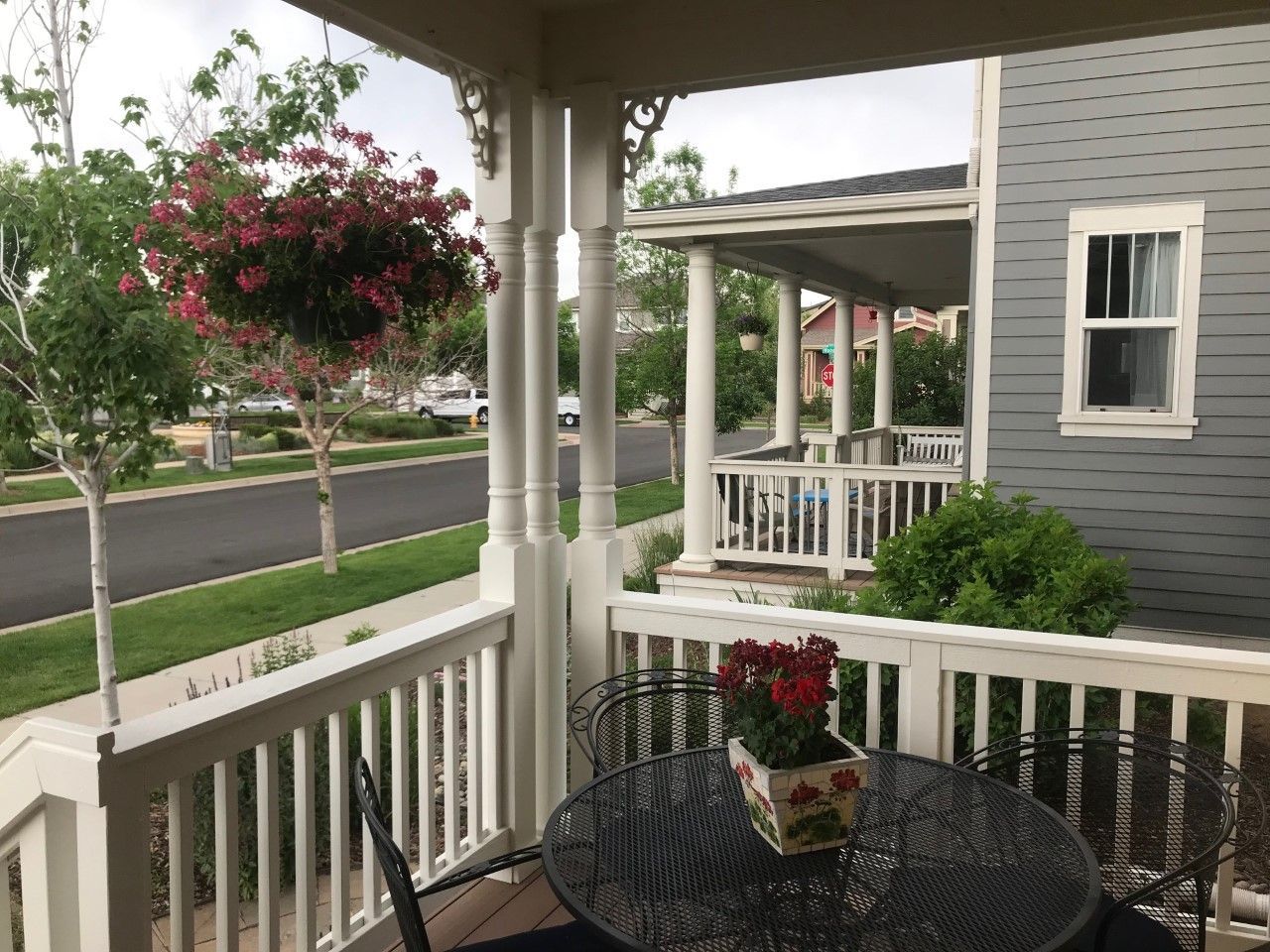 Front porch with white railing, black table, hanging flowers, and a quiet suburban street view.