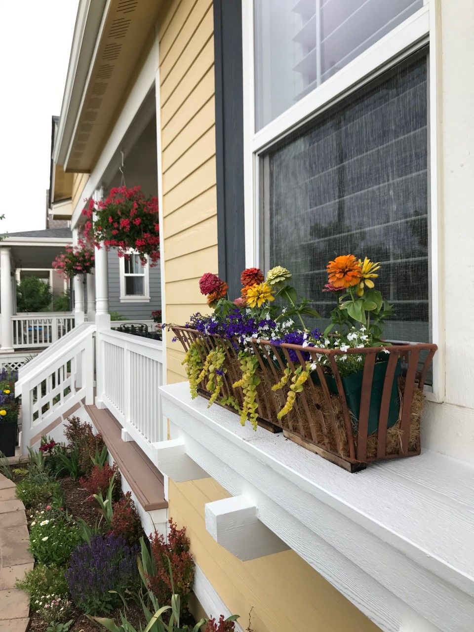 Yellow house with flower box on window sill, porch with hanging baskets.
