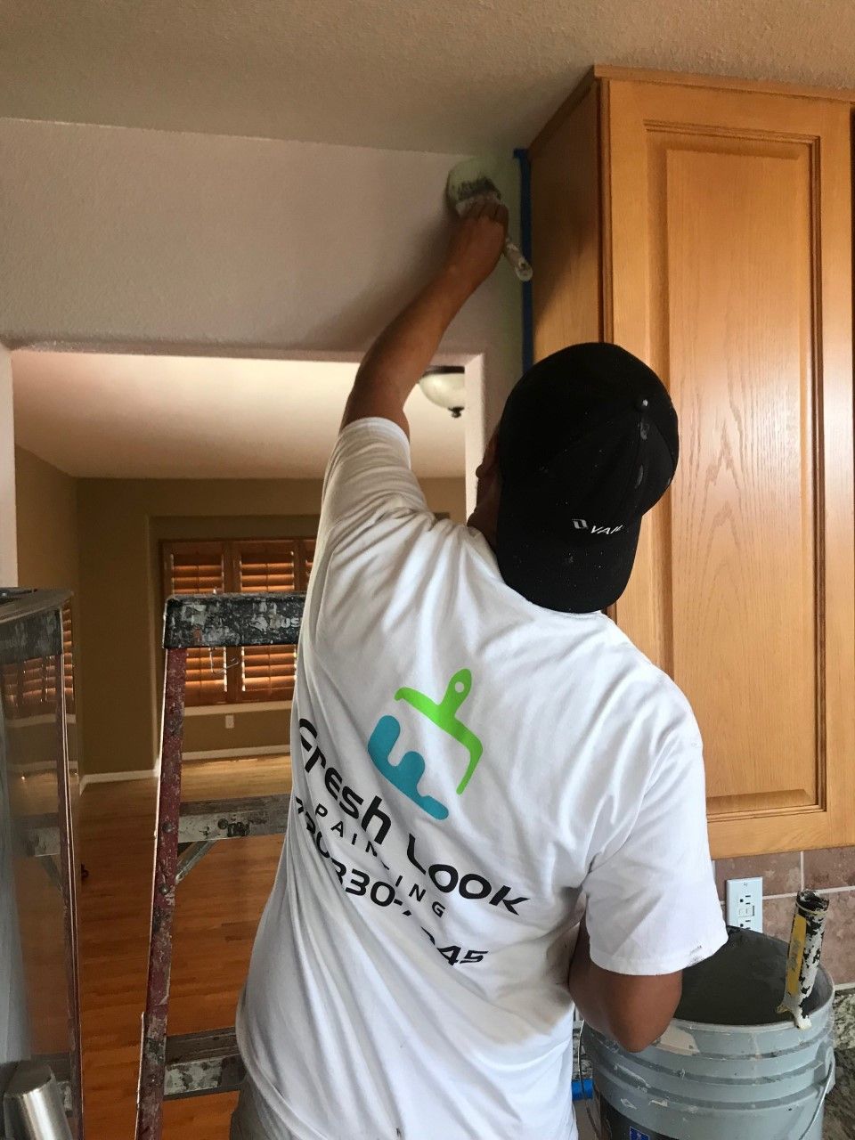Worker painting a wall above kitchen cabinets with a roller, reaching up on a ladder.