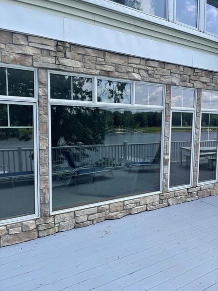 Large stone-trimmed windows on a house exterior, reflecting a lake and deck railing