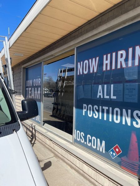 Storefront window with large “NOW HIRING ALL POSITIONS” sign and Domino’s logo