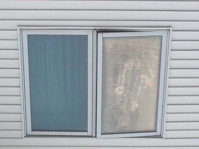 Two sliding windows on a white siding wall, one blue-tinted and one covered with a sheer curtain.