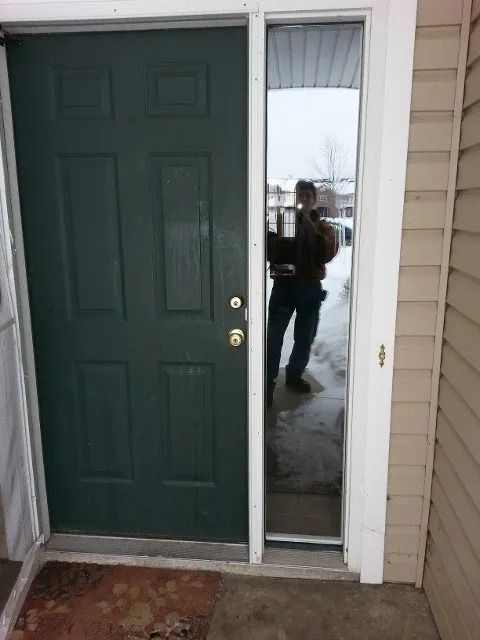 Green front door with a narrow side window, showing a person reflected in the glass.
