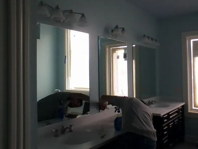 Person cleaning a bathroom sink in a blue-green bathroom with twin mirrors and wall lights