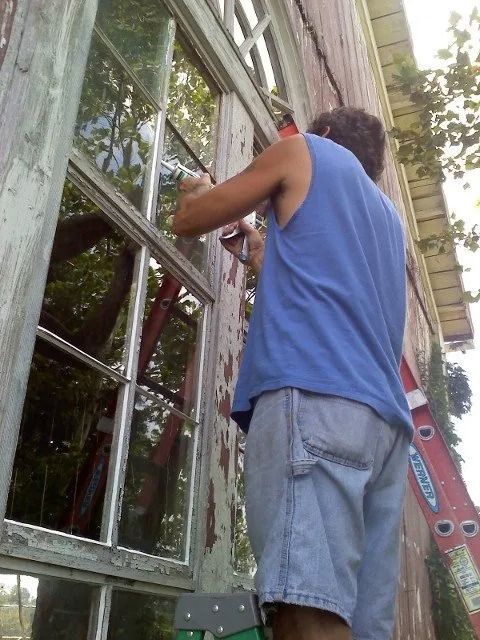 Person painting a window frame on a ladder beside a house, with a blue ladder nearby.