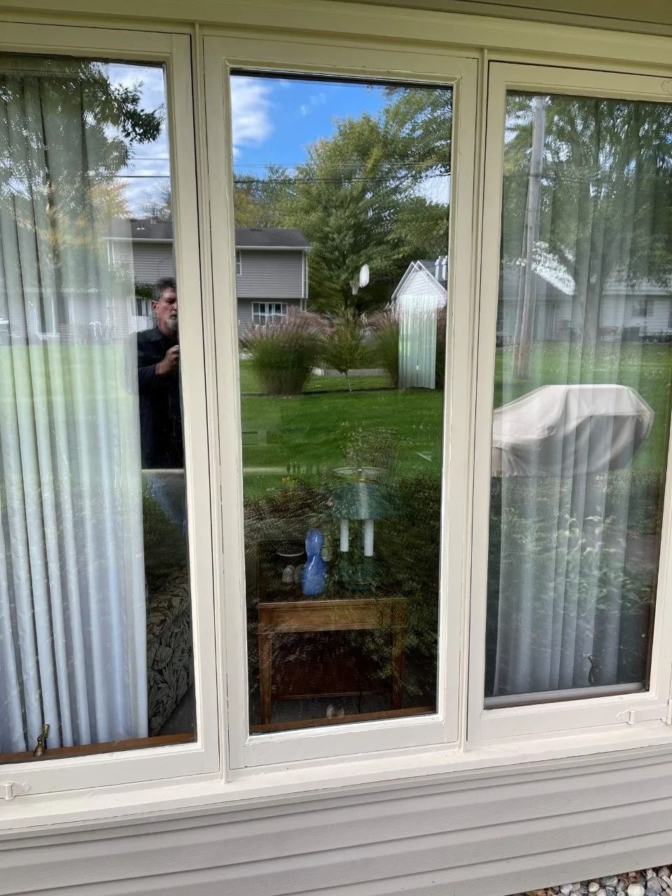 White-framed window reflecting a backyard with trees, a shed, and a person taking the photo