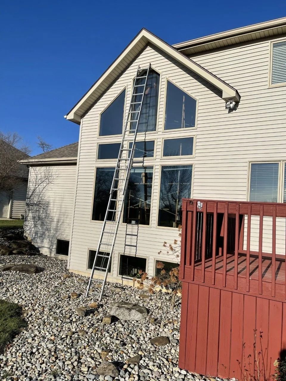 House exterior with tall ladder against windows and a red deck, surrounded by gravel under a blue sky