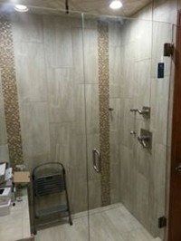 Small tiled shower with glass door, wall-mounted controls, and a folding chair inside.
