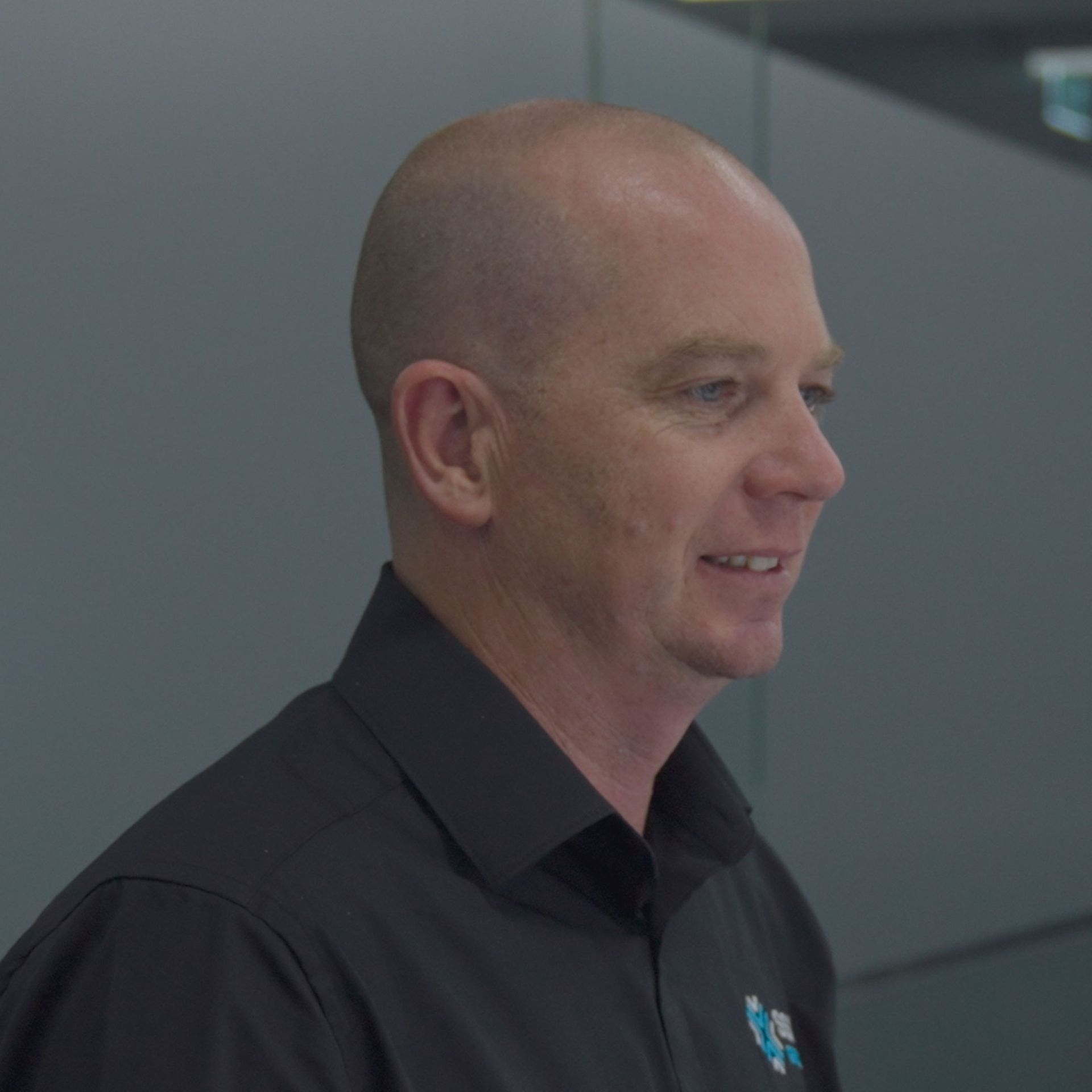 A bald man wearing a black shirt poses in front of a glass wall, reflecting a modern and professional setting.