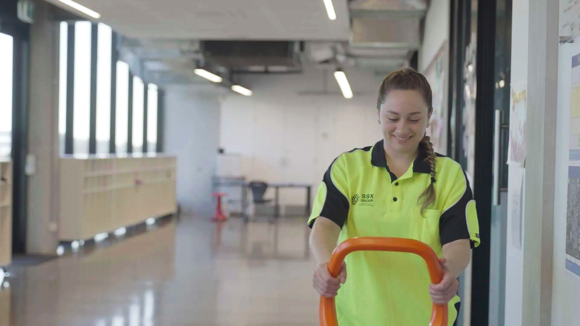 A woman wearing a yellow shirt and safety vest is seen cleaning a hallway, showcasing the efforts of the SSX group.  