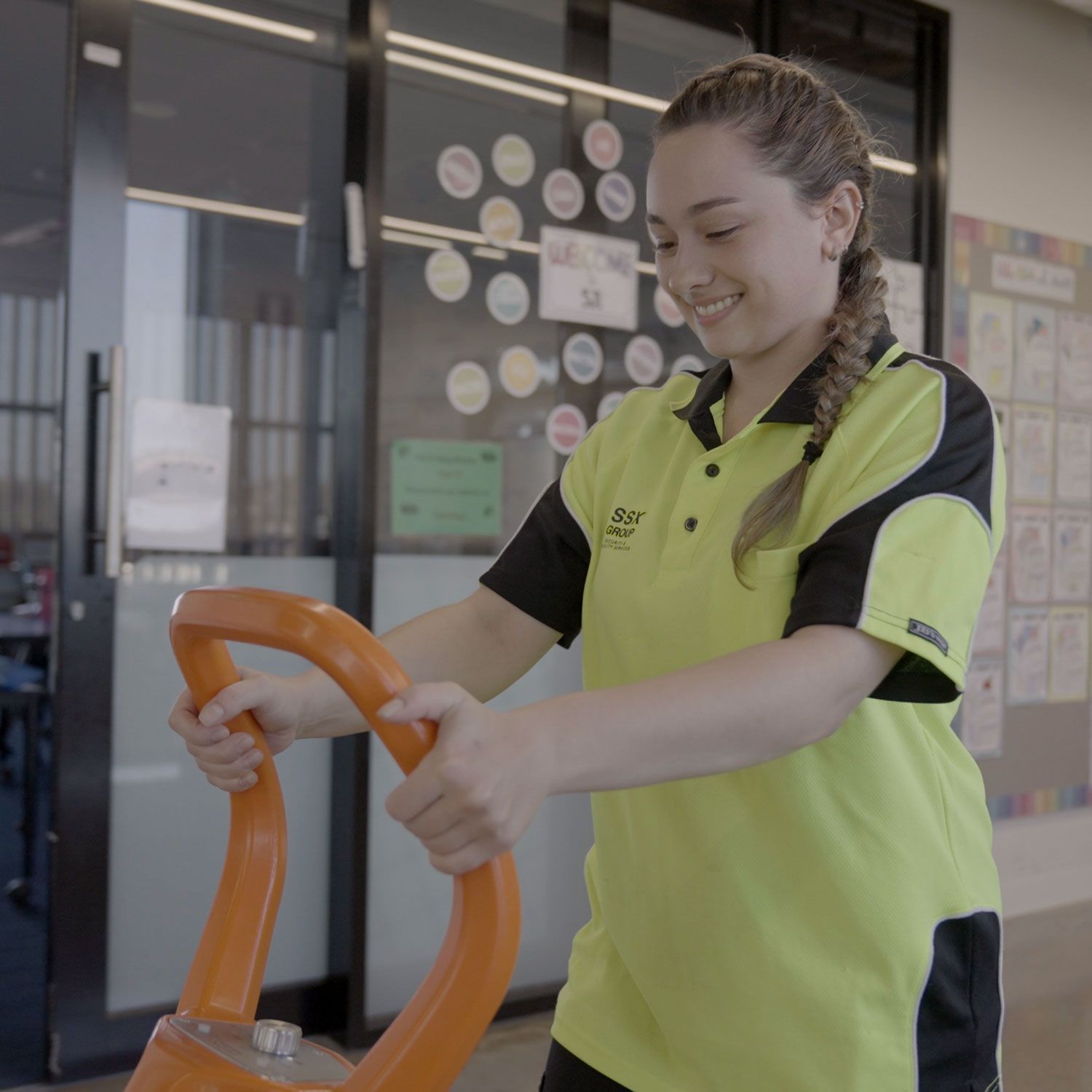 A woman in a yellow shirt from cleaning services is pushing an orange item