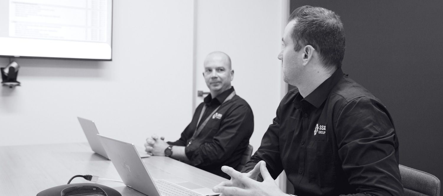 Two men working on laptops at a table in a conference room, focused on their tasks for SSX group facility maintenance.