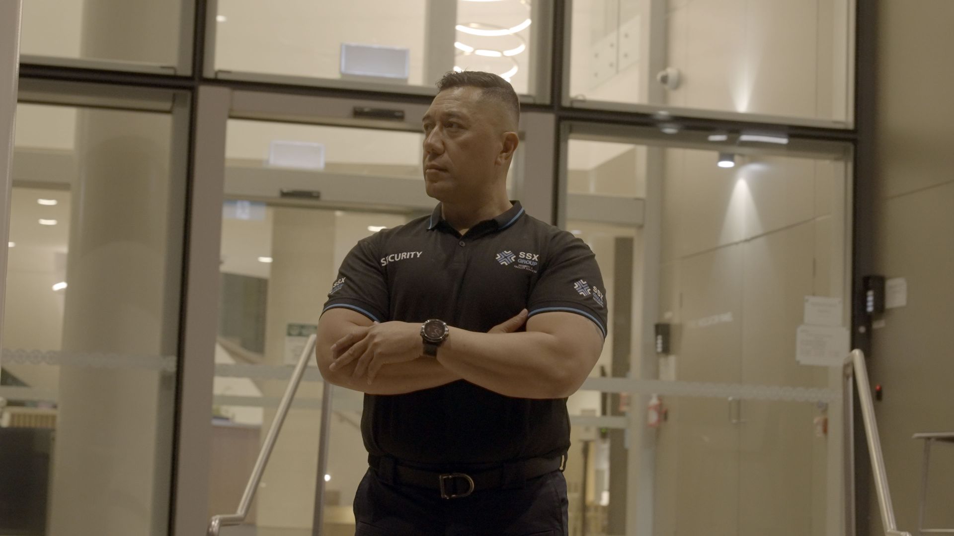 A SSX group security guard is standing in front of a glass door with his arms crossed.
