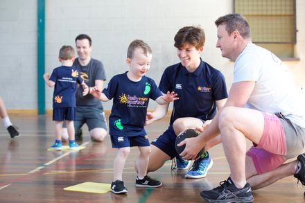 Children and adults kneel on a gym floor, cheering during a playful indoor activity.
